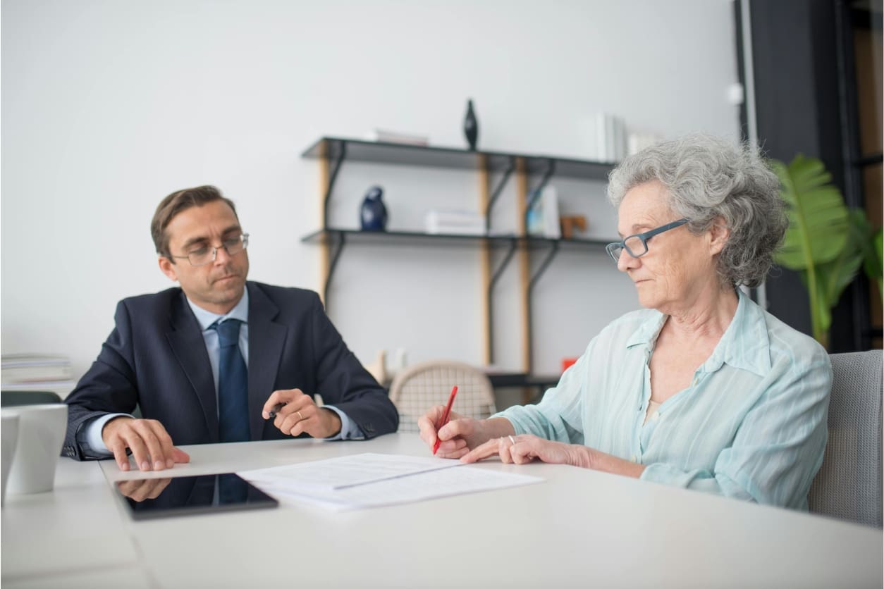 Senior woman signing documents while a man attentively watches.